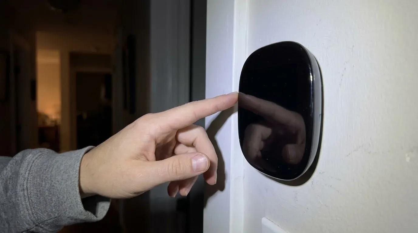 A finger touching a black smart thermostat on a white hallway wall at night, its screen completely dark